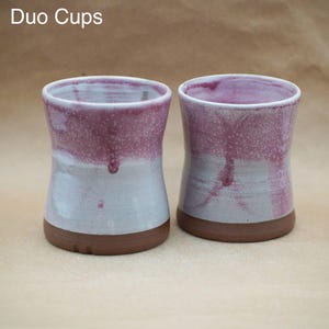 May include: Two ceramic cups labeled Duo Cups, featuring a cylindrical shape with a slight inward curve. The upper portion has a speckled magenta glaze that drips down over a light grey base, while the bottom rim remains exposed brown clay.