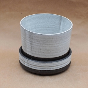 May include: A cylindrical ceramic planter with a speckled white glaze on the upper portion and a solid dark charcoal base. The planter features a matching circular saucer at the bottom with horizontal ridged detailing around the exterior rim.