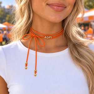 Può includere: Una collana girocollo in cordino arancione con le lettere ADPI in perline dorate. Il cordino si avvolge attorno al collo e si annoda lateralmente, con due estremità pendenti rifinite da perline sferiche dorate e piccoli ciondoli a cuore.