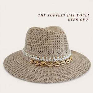 May include: A beige fedora-style hat with a wide brim. The hat features a decorative band of white pearls, gold beads, and gold-colored seashells. The text "THE SOFTEST HAT YOULL EVER OWN" is printed on the image.