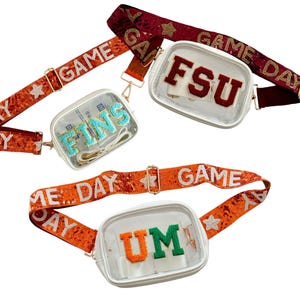 May include: Two clear plastic crossbody bags with adjustable straps. The bags have white frames and feature embroidered letters. One bag has the letters "FSU" in maroon and the other has the letters "UM" in orange and green. The straps are decorated with sequins and the words "Game Day".