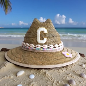 May include: A tan straw fedora hat featuring a white letter C patch on the front, a band of small white cowrie shells, a thin strand of multicoloured beads, and a single pink and white flower applique on the wide brim with a light trim.