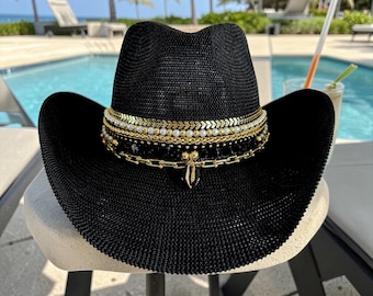Puede incluir: Un sombrero de vaquero de paja negra con una banda decorativa de varias capas que incluye eslabones dorados en forma de cheurón, pequeñas perlas blancas, cuentas doradas, cuentas negras facetadas y una cadena dorada con un colgante central negro.