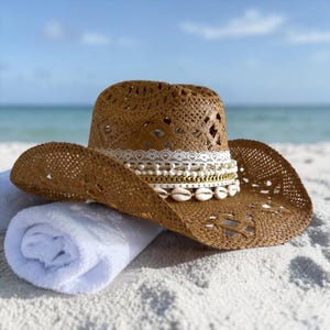 Könnte beinhalten: Ein brauner Cowboyhut aus gewebtem Stroh mit einem dekorativen Band aus weißer Spitze, goldenen Ketten, Perlen und Muscheln. Der Hut liegt auf einem gerollten weißen Handtuch an einem Sandstrand, mit dem Meer im Hintergrund.