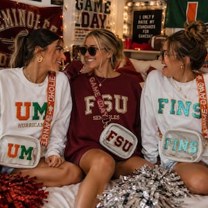 May include: Three long-sleeve sweatshirts in white and maroon featuring sequined lettering that reads UM, FSU, and FINS. Each sweatshirt is paired with a clear crossbody bag with matching sequined text and a decorative strap labeled GAME DAY.