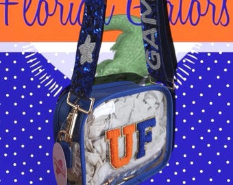 May include: A blue and clear crossbody bag with the letters "UF" in orange and blue. The bag has a blue strap with silver star and sequin details. The background is orange and blue with the words "Florida Gators".