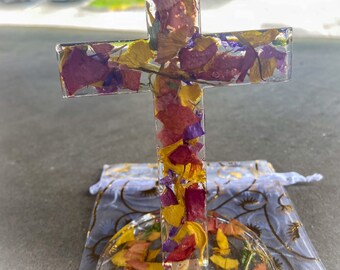 May include: A clear resin cross filled with dried flowers in shades of yellow, purple, and red. The cross is mounted on a clear, circular base, and rests on a white and gold patterned fabric.