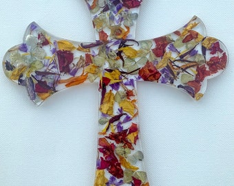May include: A clear resin cross filled with dried flower petals in shades of red, yellow, purple, and white. The cross has a decorative, ornate shape with flared ends.