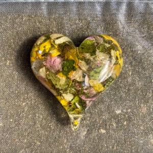 May include: A heart-shaped resin pendant filled with dried flowers in shades of yellow, pink, and green. The clear resin allows the flowers to be visible. The pendant has a pointed bottom.
