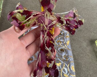 May include: A clear resin cross filled with dried burgundy and purple flower petals and gold flakes. The cross is held over a gray and gold patterned fabric.