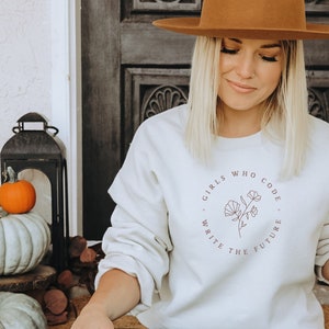 May include: A cream-coloured sweatshirt with the text "GIRLS WHO CODE WRITE THE FUTURE" in a circular design. The person is holding a white coffee cup with a green logo. The person is wearing a brown hat and black jeans.