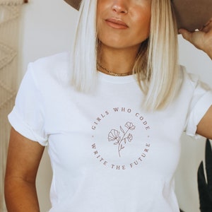 May include: White t-shirt with rolled sleeves featuring a circular design with the text "GIRLS WHO CODE WRITE THE FUTURE" and a floral illustration in a muted red-brown color. The shirt is worn with blue jeans.