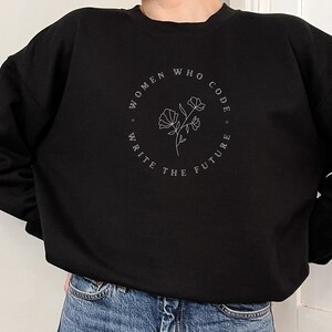 Könnte beinhalten: Schwarzes Sweatshirt mit dem Text "WOMEN WHO CODE. WRITE THE FUTURE" in einem kreisförmigen Design mit einer floralen Illustration. Das Sweatshirt hat eine schwarze Farbe.