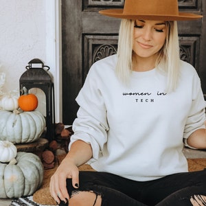 Könnte beinhalten: Ein cremefarbenes Sweatshirt mit dem Text "women in TECH" in handschriftlicher Schrift. Die Person hält einen Starbucks-Kaffeebecher. Der Hintergrund umfasst Kürbisse und Herbstdekoration.