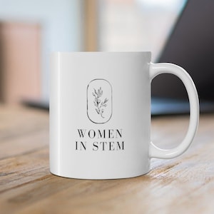 Puede incluir: Taza de cerámica blanca con un diseño floral negro dentro de un óvalo y las palabras "WOMEN IN STEM" impresas en negro. La taza está sobre una superficie de madera, con un portátil en el fondo.