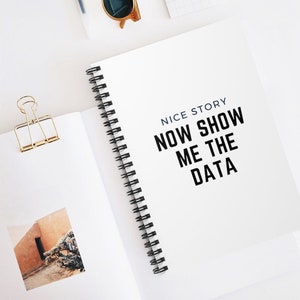 May include: A white spiral bound notebook with the text "NICE STORY NOW SHOW ME THE DATA" printed in black on the cover.