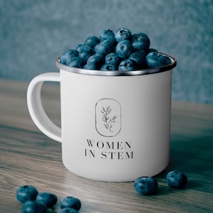 May include: White enamel mug with a black and white logo that says "Women in STEM" and a small floral design. The mug is filled with blueberries.