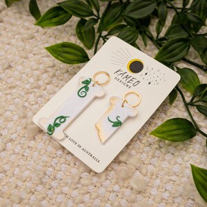 May include: Two white keychains with gold rings and green leaf designs. The keychains are on a white card with the text "KAMEO DESIGNS" and "with love in Australia".