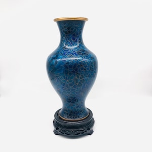 Peut inclure: Un vase cloisonné bleu orné d'un motif floral détaillé souligné par de fins fils dorés. Le vase présente un col étroit, un bord évasé avec une finition dorée et un corps bombé. Il repose sur un socle circulaire en bois sombre sculpté.
