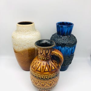 Puede incluir: Tres jarrones de cerámica de diferentes tamaños y colores. El jarrón más alto es beige y marrón, el jarrón del medio es marrón con asa y el tercer jarrón es azul y negro. Los jarrones tienen superficies texturizadas y diseños decorativos.