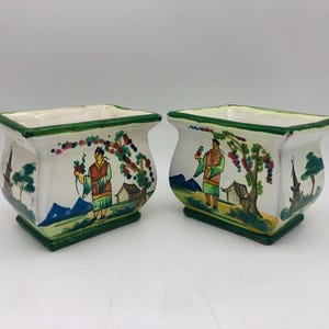 Peut inclure: Deux jardinières rectangulaires en céramique avec une base blanche et une bordure verte. Chaque jardinière présente des illustrations peintes à la main de personnages en vêtements traditionnels, d'arbres et de petits bâtiments.