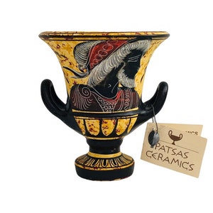 May include: A black and gold ceramic vase with two handles, featuring a painted illustration of a figure in profile. The vase has a decorative design and the text "PATSAS CERAMICS" on a tag.