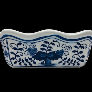Peut inclure: Un plat rectangulaire en céramique avec un bord supérieur ondulé, présentant une base blanche ornée d'un motif floral et végétal bleu. Le design comprend des feuilles stylisées, des tiges en spirale et des motifs en forme de nuages.