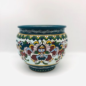 以下が含まれることがあります： 丸いセラミック製の植木鉢で、縁と底はティール色です。植木鉢の本体は水色で、赤、黄、青、緑の色合いの花柄模様が施されています。植木鉢には金のトリムが付いています。