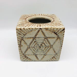 Op de afbeelding: Een vierkante, gebroken witte keramische tissueboxhouder met een geometrisch ontwerp. De bovenkant heeft een cirkelvormige opening omgeven door radiale lijnen. De zijkanten hebben een zespuntig sterpatroon met geëtste lijnen.