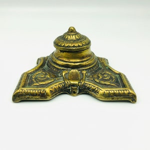May include: A brass inkwell featuring a wide, triangular base with ornate, raised scrollwork patterns and a central, domed lid. The metal surface displays a weathered, aged patina with dark recessed areas and polished highlights on the edges.