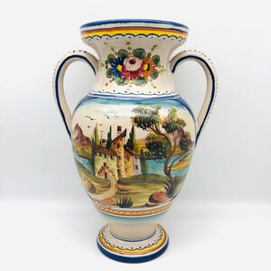 May include: A hand-painted ceramic vase with two handles. The vase features a landscape scene with buildings, trees, and a body of water. The top rim and base have blue and yellow accents. Floral designs adorn the top of the vase.
