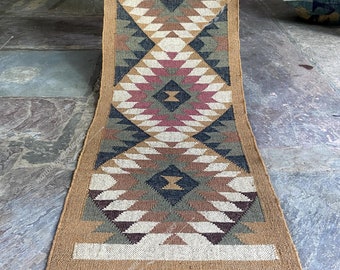 May include: A brown and beige jute rug with a geometric pattern in shades of brown, beige, white, black, and pink. The rug has a fringe border.