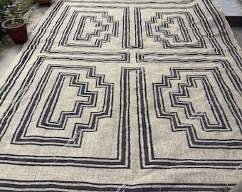 May include: Large rectangular rug with a geometric pattern. The rug features a beige background with a repeating design of dark gray lines forming rectangular and stepped shapes. The rug is made of a woven material.