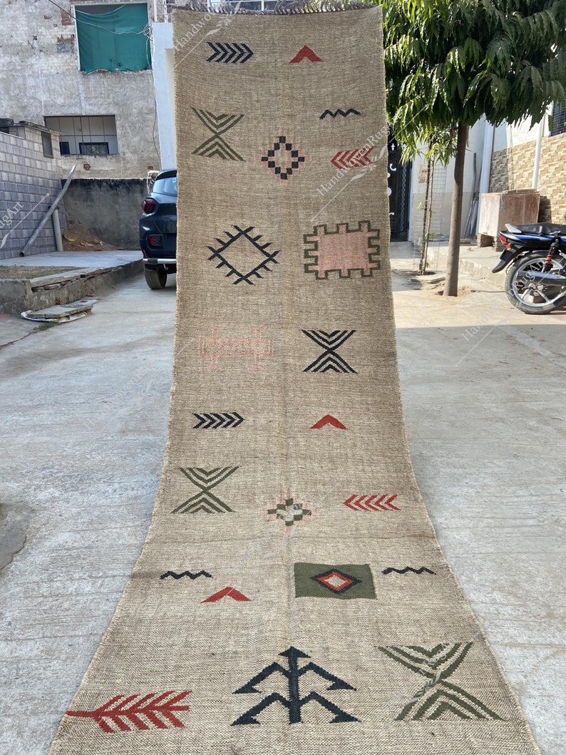 May include: A long, beige runner rug with geometric patterns in black, red, green, and pink. The rug features various shapes, including arrows, diamonds, and zigzags, creating a textured, decorative design. The rug is displayed outdoors.