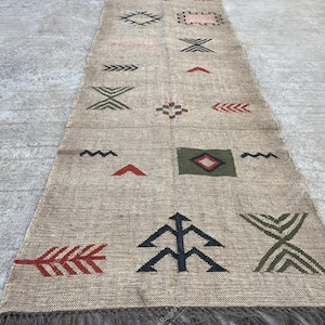 May include: A beige runner rug with geometric patterns in red, green, black, and pink. The rug features a fringed edge and various shapes, including arrows, diamonds, and zigzags. The rug is made of a woven material.