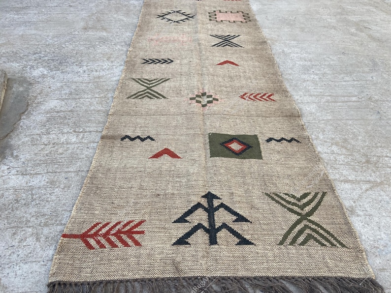 May include: A beige runner rug with geometric patterns in red, green, black, and pink. The rug features a fringed edge and various shapes, including arrows, diamonds, and zigzags. The rug is made of a woven material.