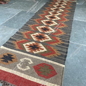 Puede incluir: Una alfombra larga y estrecha con un patrón geométrico en tonos marrones, rojos y blancos. La alfombra tiene flecos en un extremo.