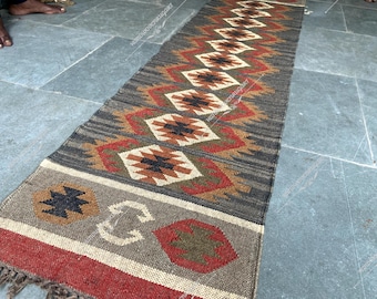May include: A long, narrow rug with a geometric pattern in shades of brown, red, and white. The rug has a fringe on one end.