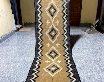 May include: A long, narrow rug with a geometric pattern in black, white, and beige. The rug has a fringe on the ends.