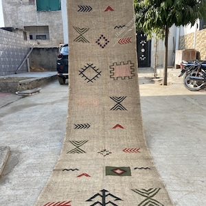 May include: A long, rectangular beige rug with a geometric pattern in black, red, and green. The rug features various shapes, including arrows, diamonds, and rectangles. The edges have a fringe detail.