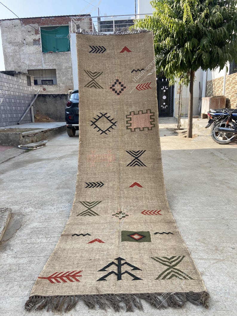 May include: A long, rectangular beige rug with a geometric pattern in black, red, and green. The rug features various shapes, including arrows, diamonds, and rectangles. The edges have a fringe detail.