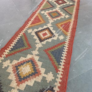 May include: A colorful, geometric patterned rug with a red, green, blue, and beige color scheme. The rug is made of woven jute and has a rustic, hand-crafted look.