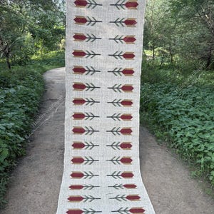 Puede incluir: Una alfombra rectangular de color blanquecino con un patrón repetido de formas rojas y doradas en forma de flecha con detalles en verde. La alfombra tiene un borde con flecos y se exhibe al aire libre en un camino.