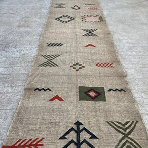 May include: A long, rectangular beige rug with geometric patterns in black, red, and green. The rug features various shapes, including arrows, diamonds, and zigzags, creating a textured, decorative design. The rug has a fringed edge.