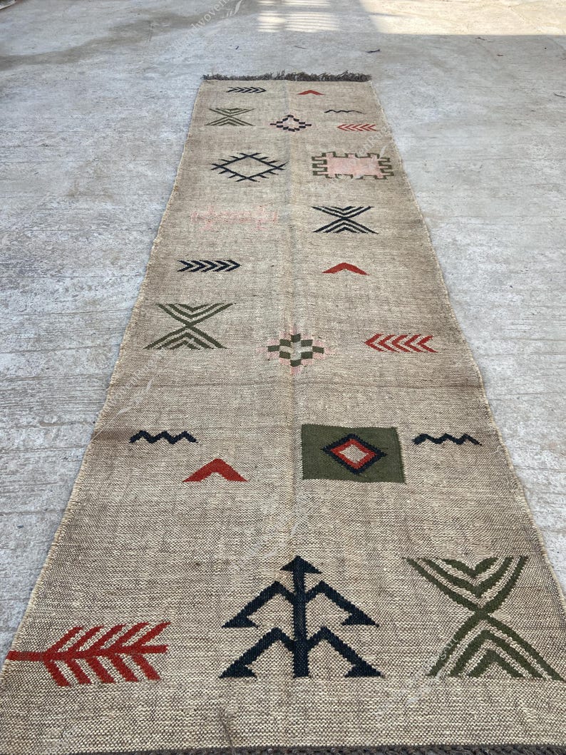 May include: A long, rectangular beige rug with geometric patterns in black, red, and green. The rug features various shapes, including arrows, diamonds, and zigzags, creating a textured, decorative design. The rug has a fringed edge.