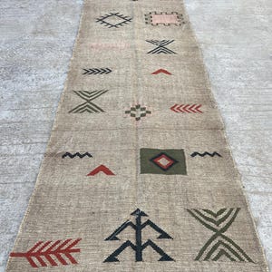 May include: Beige runner rug with geometric patterns in black, green, and red. The rug has a fringe on both ends and features various shapes, including arrows, diamonds, and zigzags. The rug is made of a woven material.