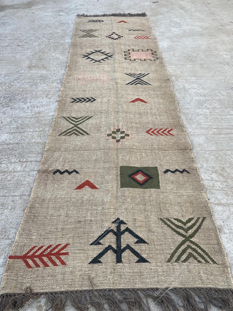 May include: Beige runner rug with geometric patterns in black, green, and red. The rug has a fringe on both ends and features various shapes, including arrows, diamonds, and zigzags. The rug is made of a woven material.