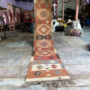 May include: A long, narrow rug with a geometric pattern in shades of red, orange, brown, and white. The rug has a fringe on both ends.