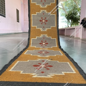 Può includere: Un tappeto lungo e stretto con un motivo geometrico in tonalità di grigio, giallo e rosso. Il tappeto ha frange su entrambi i lati.