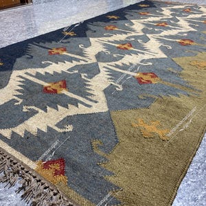 May include: A handwoven rug with a geometric pattern in shades of blue, beige, and brown. The rug has a fringe border and is made of natural fibers.